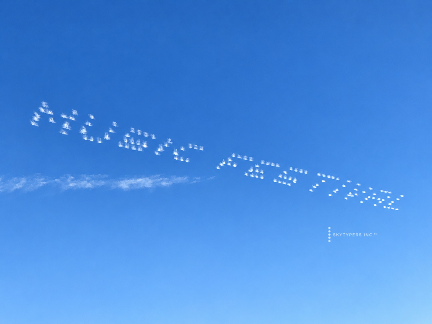 White dotted sky lettering across blue sky above a thin horizontal smoke trail.