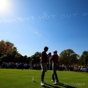 Golfers stand on a green beneath faint smoke lettering, with a crowd in the background.