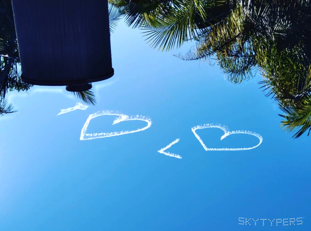 Two white smoke hearts in a vivid blue sky above palm trees.