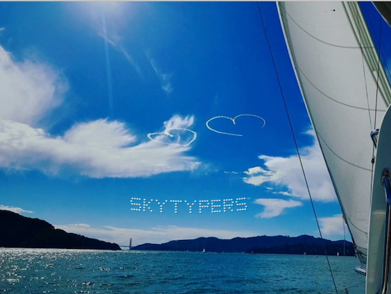 White smoke hearts above water and distant hills, with a sail at the right edge.