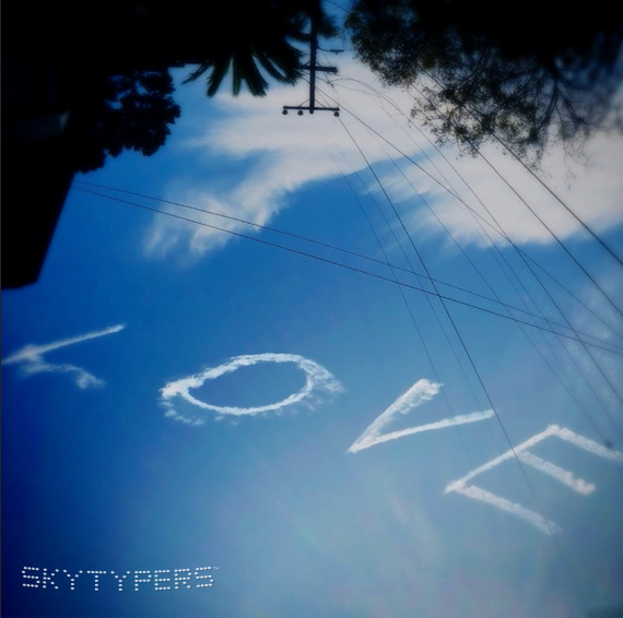 LOVE written in white smoke across a blue sky, with trees and utility wires along the top edge.
