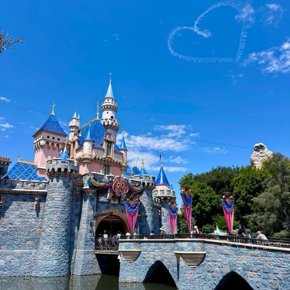 A white smoke heart above Sleeping Beauty Castle at Disneyland under a clear blue sky.