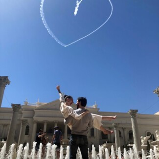A person points upward beneath a smoke heart, with a fountain and large building behind them.