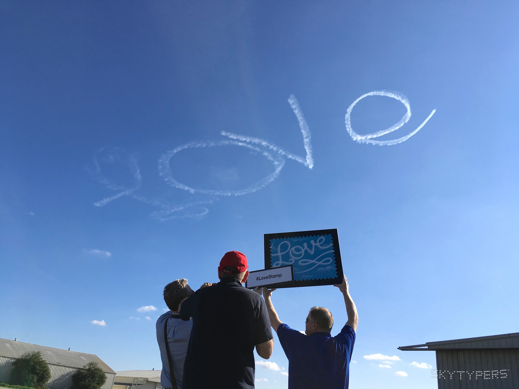 People hold a large Love stamp display beneath Love written in smoke across a blue sky.