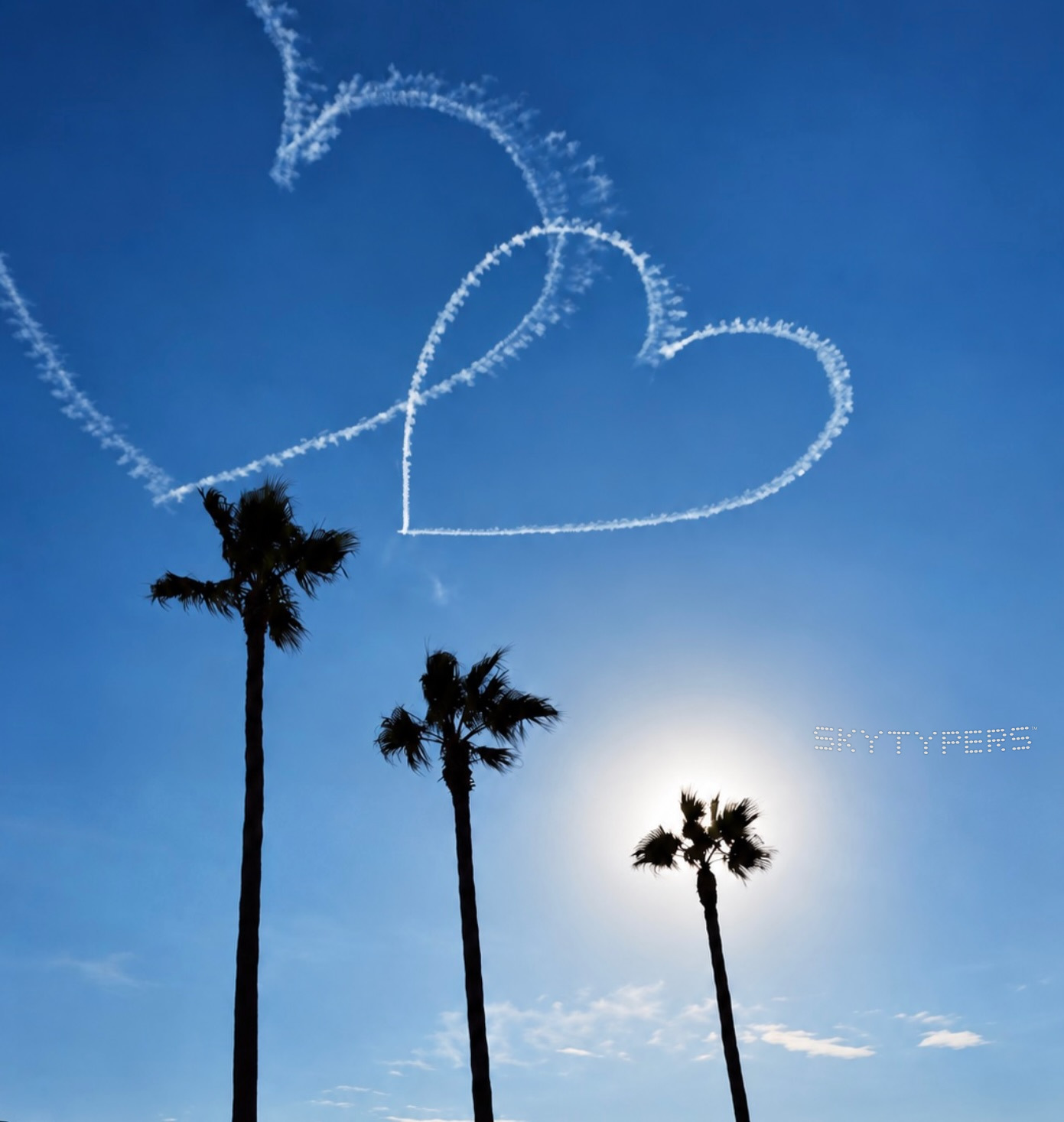 Two white smoke hearts in a blue sky above silhouetted palm trees and the sun.