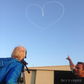 Two people gesture toward a pale heart drawn in smoke above a wall and rooftops.