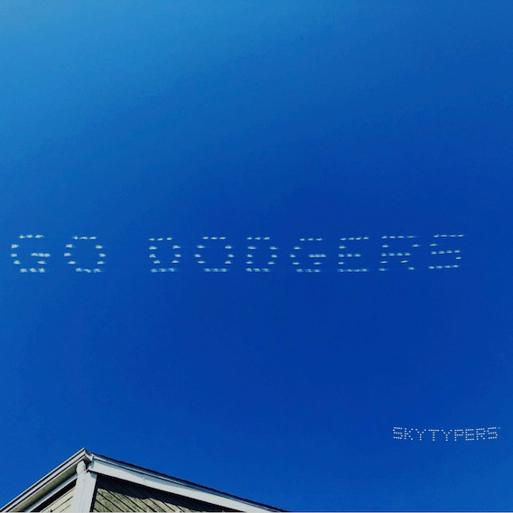 GO DODGERS skytyping across a bright blue sky, with a rooftop along the lower edge.