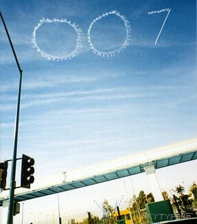 White smoke spells “007” above a street intersection, traffic signal and buildings.