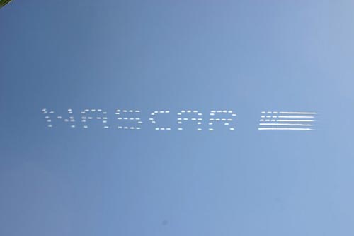 Skywriting displays the word "NASCAR" with an American flag image in a clear blue sky.