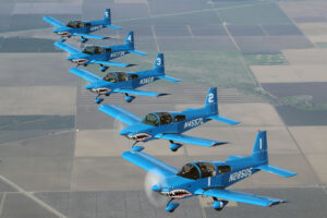 Five blue aircraft with shark mouth designs flying in formation over farmland.