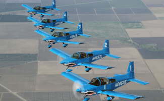 Five blue aircraft with shark mouth designs flying in formation over farmland.